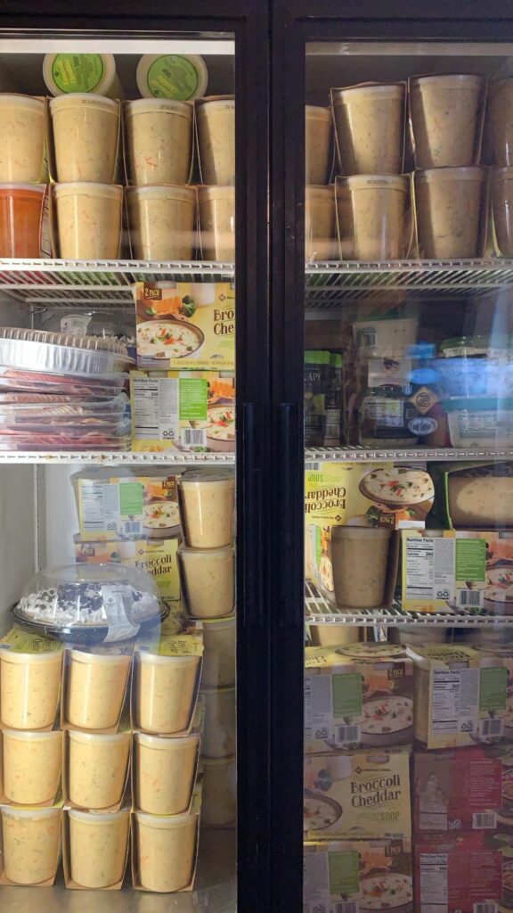 A fridge filled with stacked containers of soup and boxes of broccoli cheddar soup, assorted frozen pies, and packaged foods.