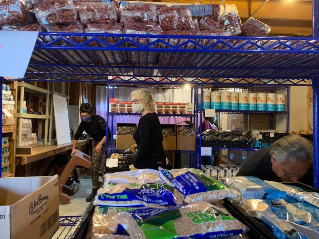 People organize items on metal shelves in a food pantry, surrounded by canned goods and bags of grains.