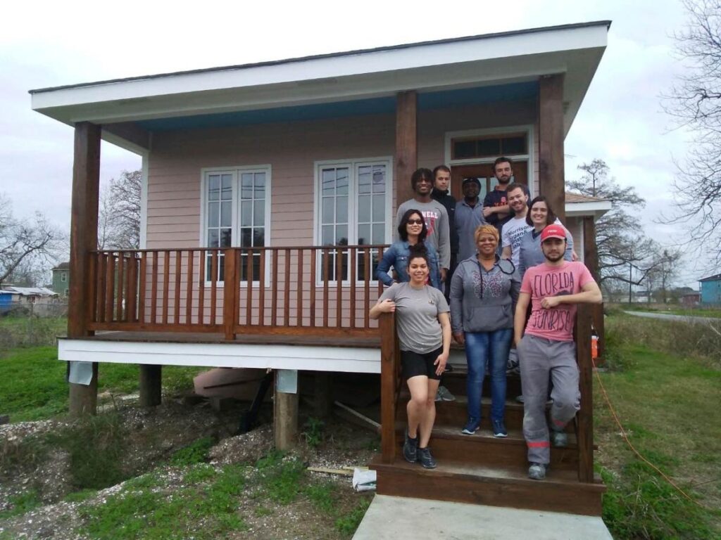 A group of ten smiling people stand on the wooden steps of a small raised house with a porch.