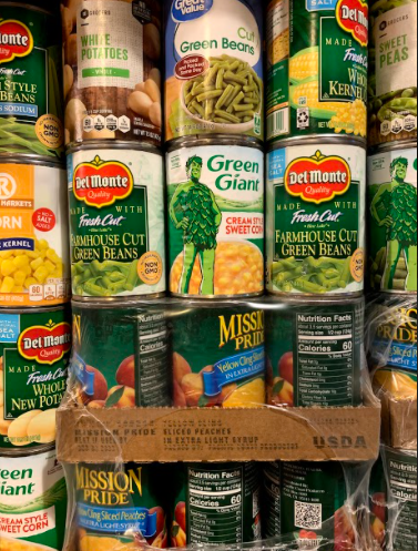 A variety of canned vegetables stacked on a shelf, including green beans, sweet peas, and peaches.