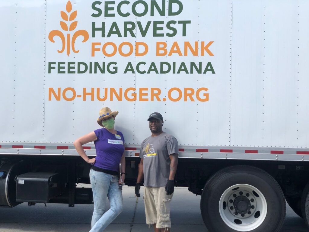 Two volunteers stand smiling in front of a Second Harvest Food Bank truck, which reads "Feeding Acadiana, No-Hunger.org."